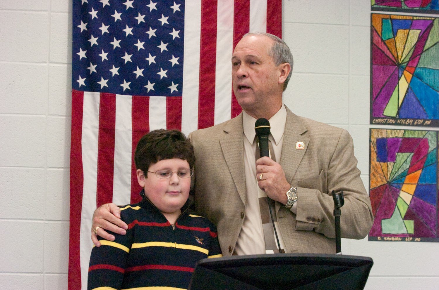 Man in a tan suit speaks into a microphone beside a seated boy, with an American flag in the background at a podium event