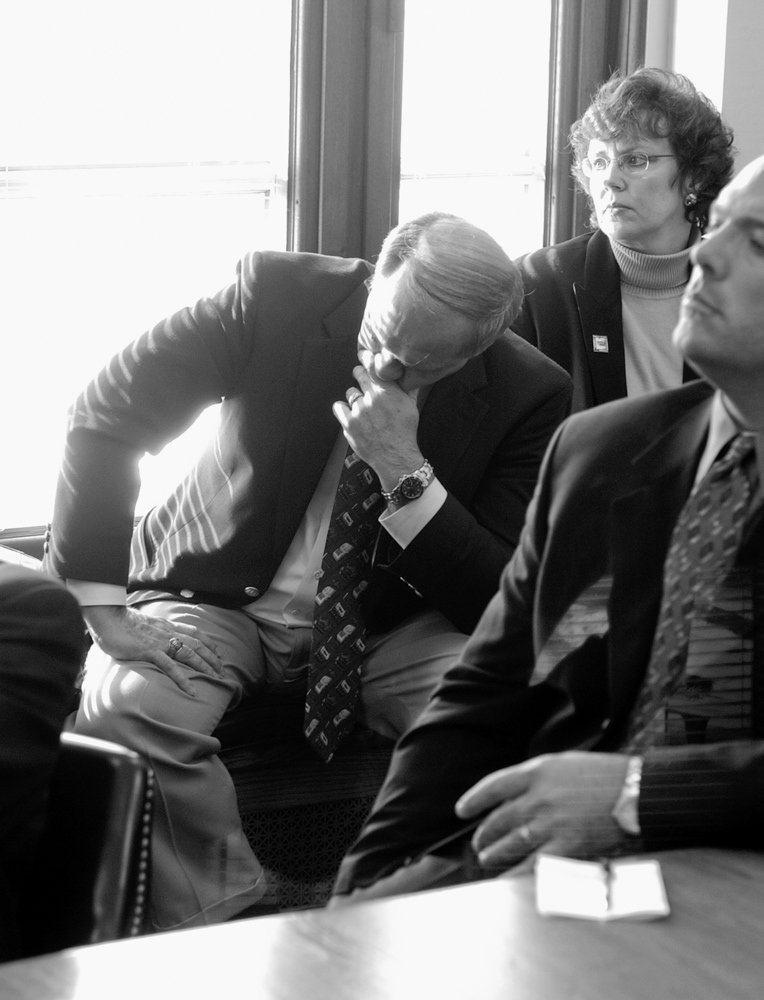 Man in a suit leans forward with his chin on his hand during a meeting, while two colleagues look on near a window.