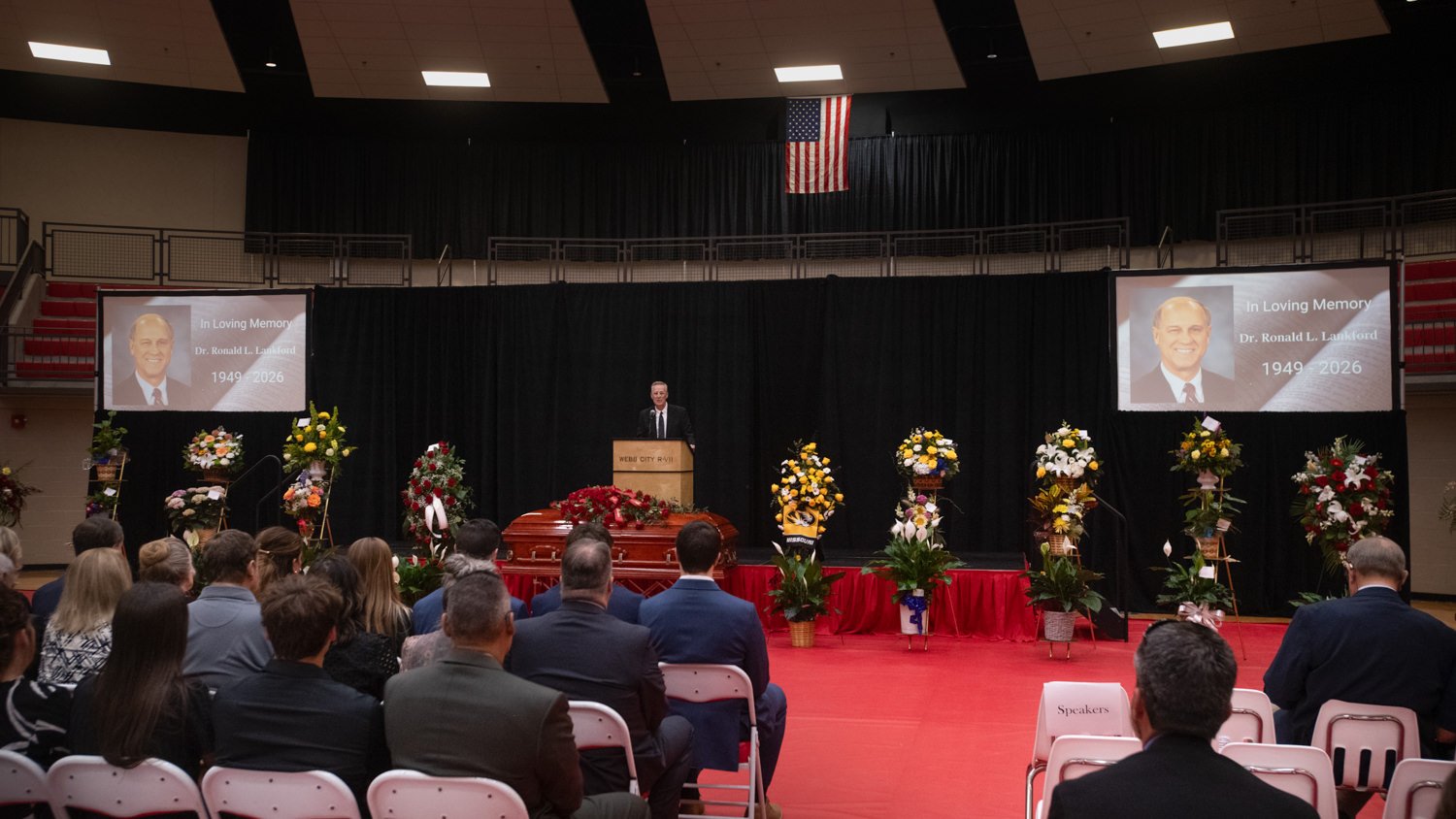 Memorial service in a gym: speaker at podium, casket on red dais, and many flower arrangements; screens honor Dr. Ronald L. Lankford (1949–2026).