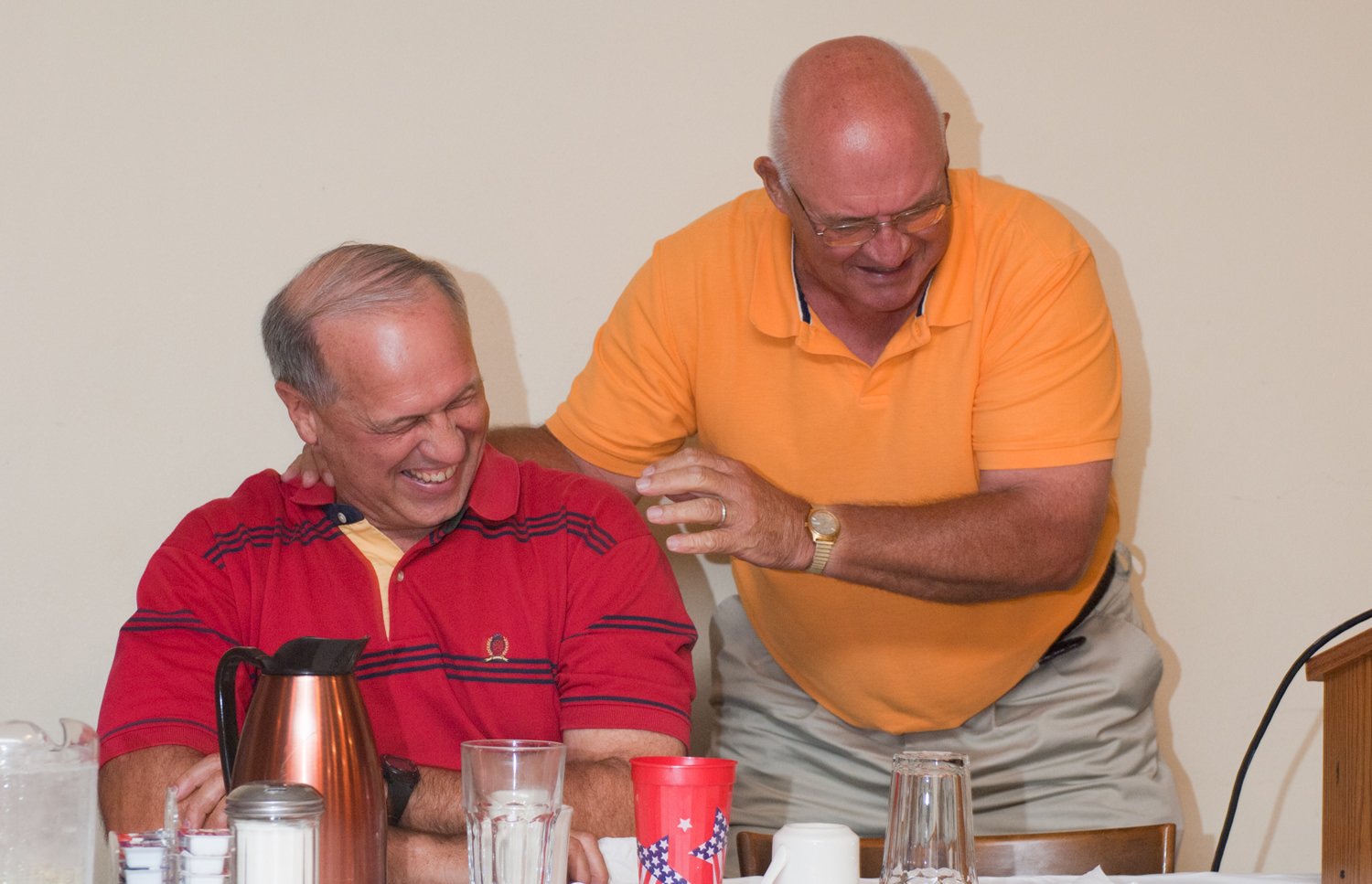 Two older men laughing together at a table, one in a red polo and the other in an orange polo, with a pitcher and cups on the table.