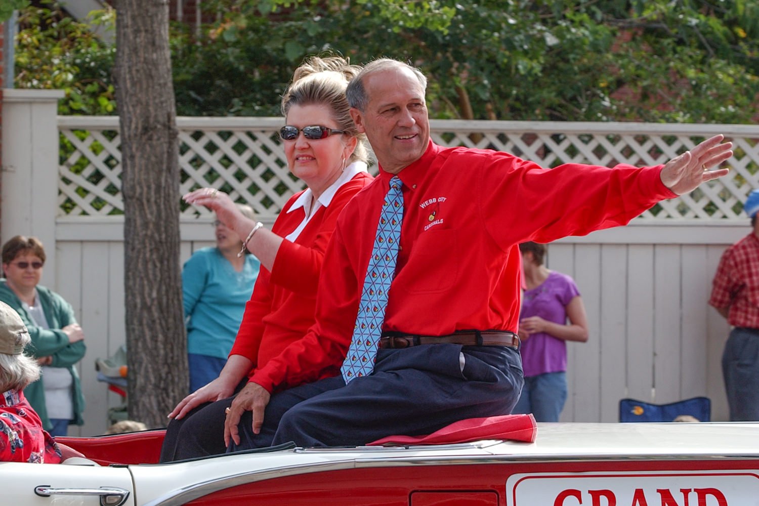 Man and woman in red shirts riding in a vintage red-and-white convertible during a parade; man extends his arms and smiles.