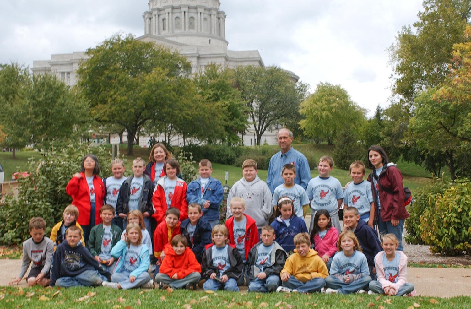 Group of children and two adults posing for a photo on a grassy lawn with a domed government building in the background.
