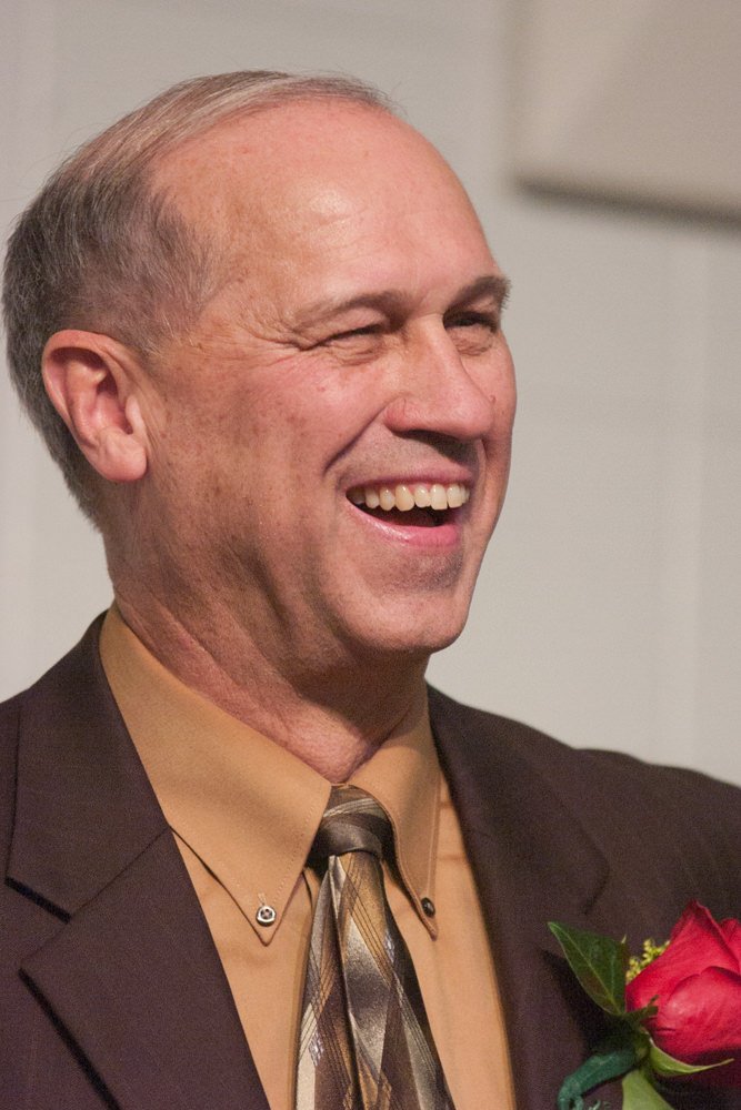 Smiling man in a brown suit with a tan shirt and striped tie, wearing a red boutonniere on the lapel.