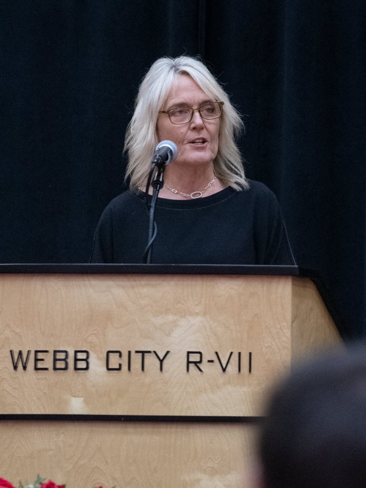 Woman with shoulder-length gray hair and glasses speaks into a microphone at a wooden podium labeled Webb City R-VII.