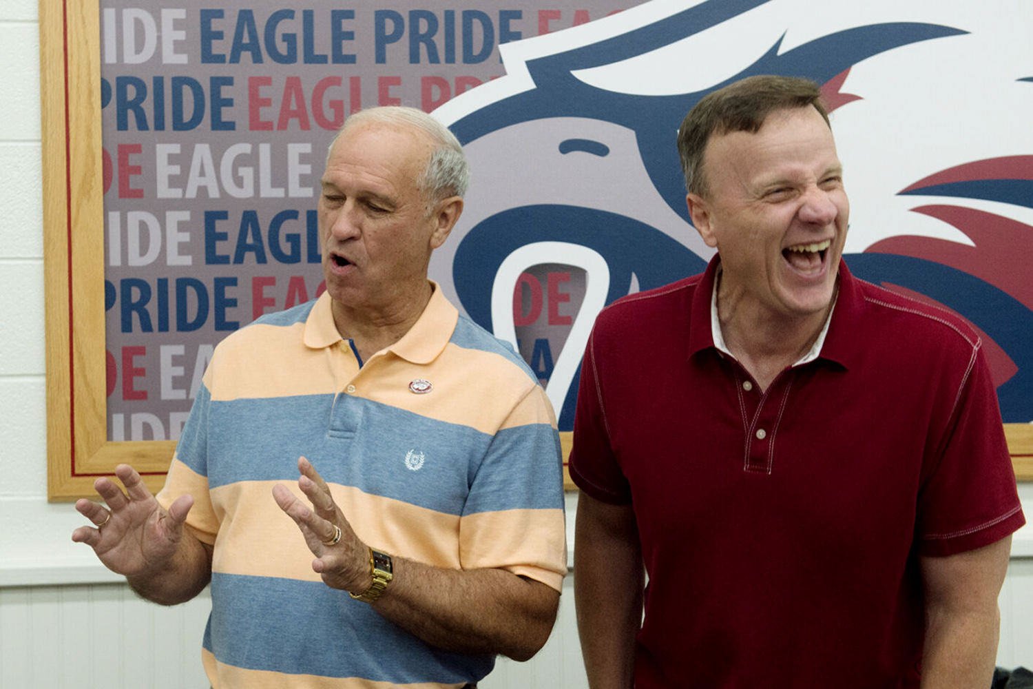 Two older men laughing together, one gesturing with his hands, in front of a large eagle logo backdrop.