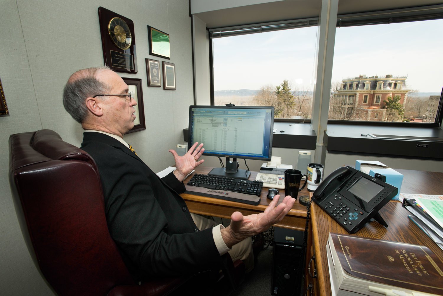 Older man in a suit sits at a cluttered office desk, gesturing with hands while looking at a computer monitor, with awards on the wall behind him and a window view outside.