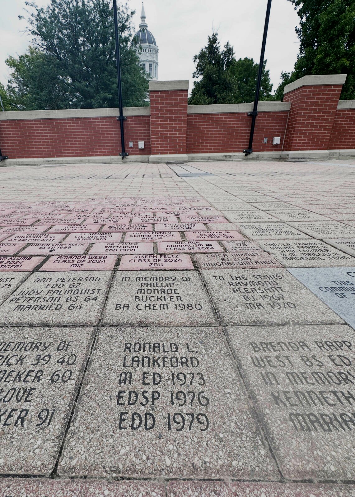 Rows of engraved stone tiles in a memorial plaza, each tile showing names and years, with a brick wall and trees in the background and a distant domed building visible.