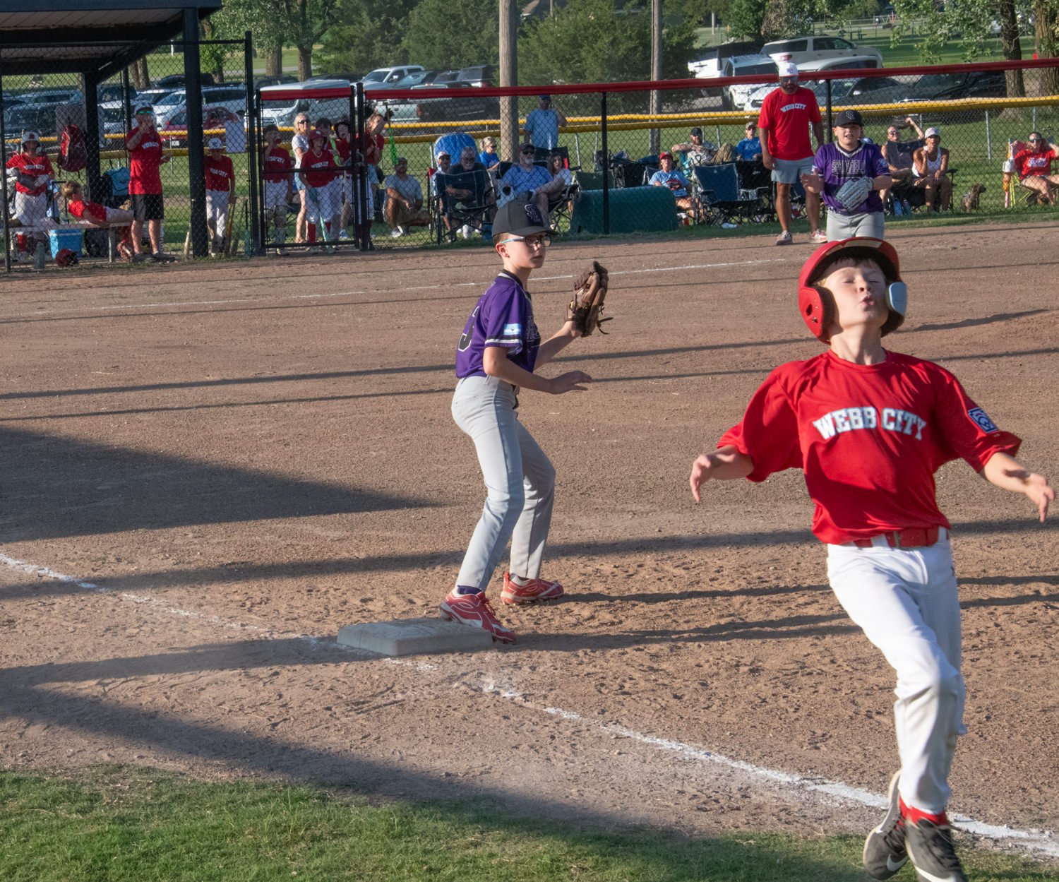 Young baseball player in a purple uniform stands on second base with glove ready, as a red-helmeted runner in a red jersey races past in the foreground.