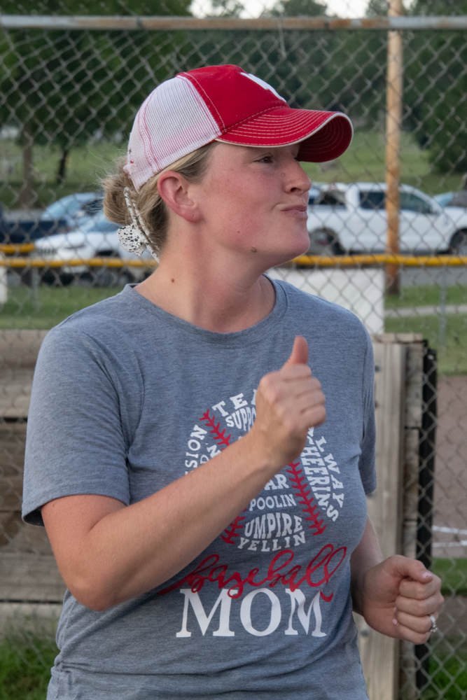 Woman in a red-and-white baseball cap and gray T-shirt with 'baseball mom' text, giving a thumbs up on a baseball field behind a chain-link fence.