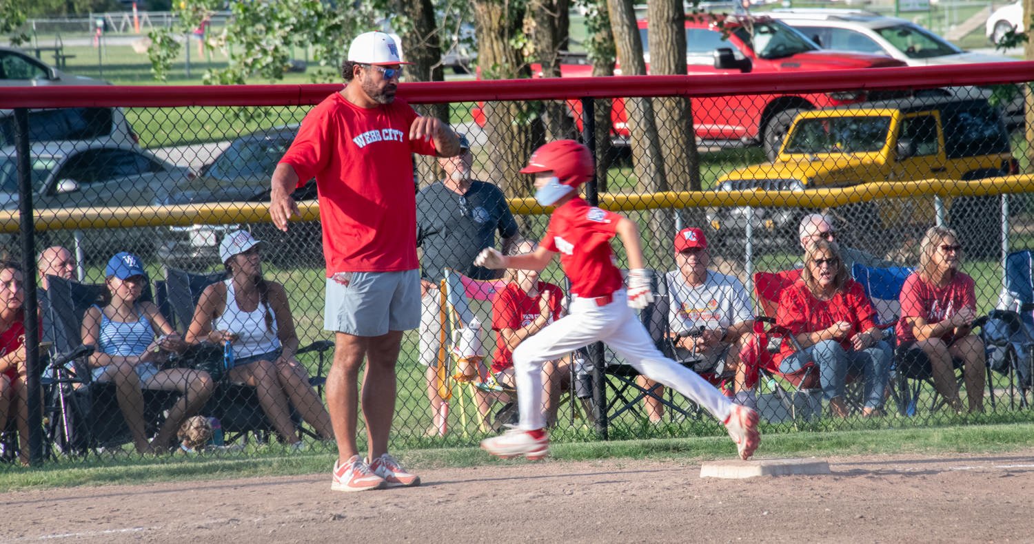 Youth baseball player in red runs toward first base as a coach in a red shirt signals nearby; spectators watch behind a chain-link fence.