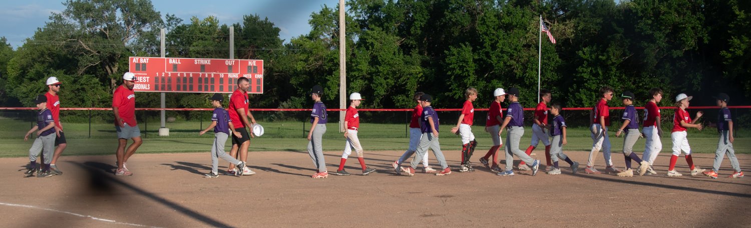 Groups of young players in red and purple uniforms line up to shake hands on a baseball field after a game, with a scoreboard and trees in the background.