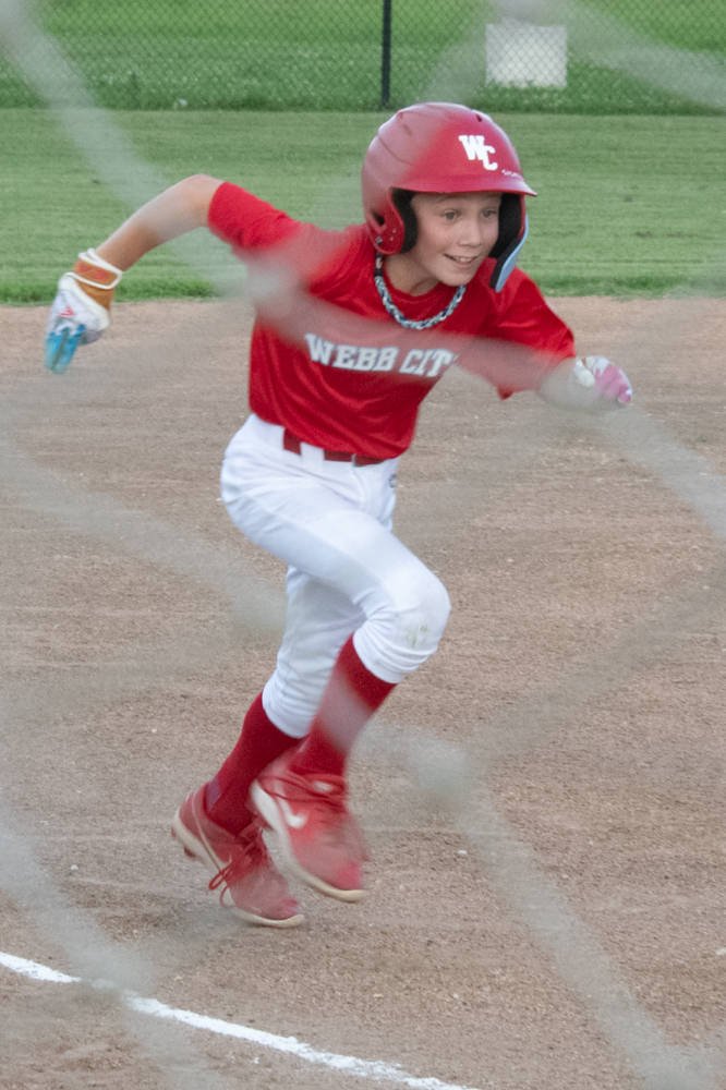 Young baseball player in a red Webb City uniform and helmet, sprinting on the dirt field near home plate.