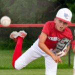 Young baseball pitcher in a red uniform throws a ball on a grassy field, with a teammate watching in the background.