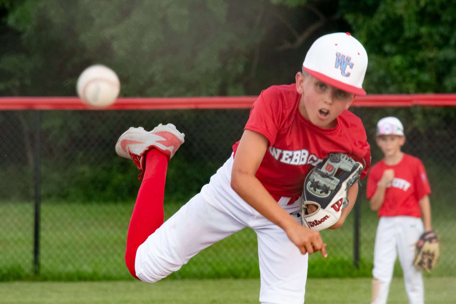 Young baseball pitcher in a red uniform throws a ball on a grassy field, with a teammate watching in the background.