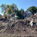 Yellow excavator at a demolition site, lifting debris with piles of rubble in front of a stone house on a sunny day