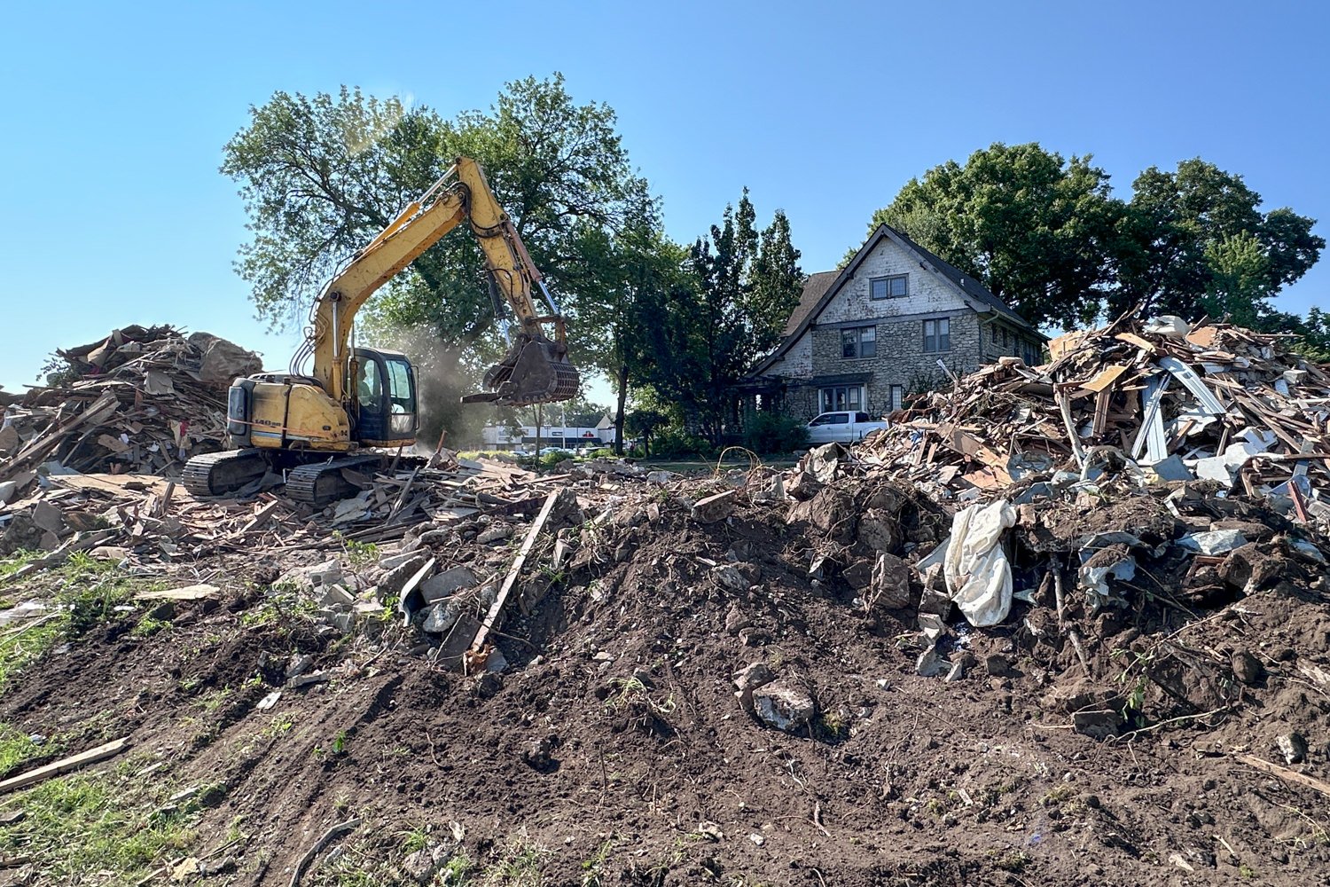 Yellow excavator at a demolition site, lifting debris with piles of rubble in front of a stone house on a sunny day