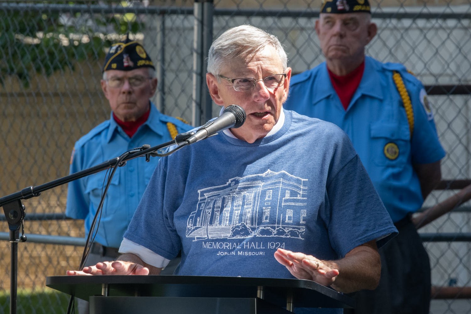 Older man speaks at a podium microphone, wearing a blue Memorial Hall 1924 shirt; two veterans stand behind him.
