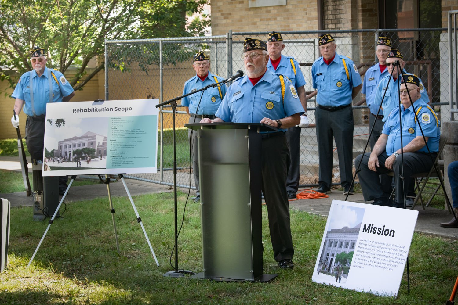 Group of veterans in blue uniforms standing behind a podium during an outdoor ceremony, with signs reading 'Rehabilitation Scope' and 'Mission' nearby.