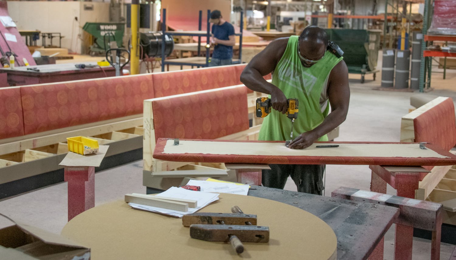 Worker in a neon-green sleeveless shirt drills a wooden board at a red workbench in a woodshop.