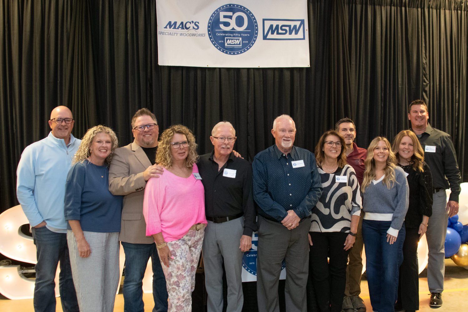 Group of about 12 adults posing for a photo at a 50th anniversary event, with a banner showing '50' and logos behind them in front of a black curtain.