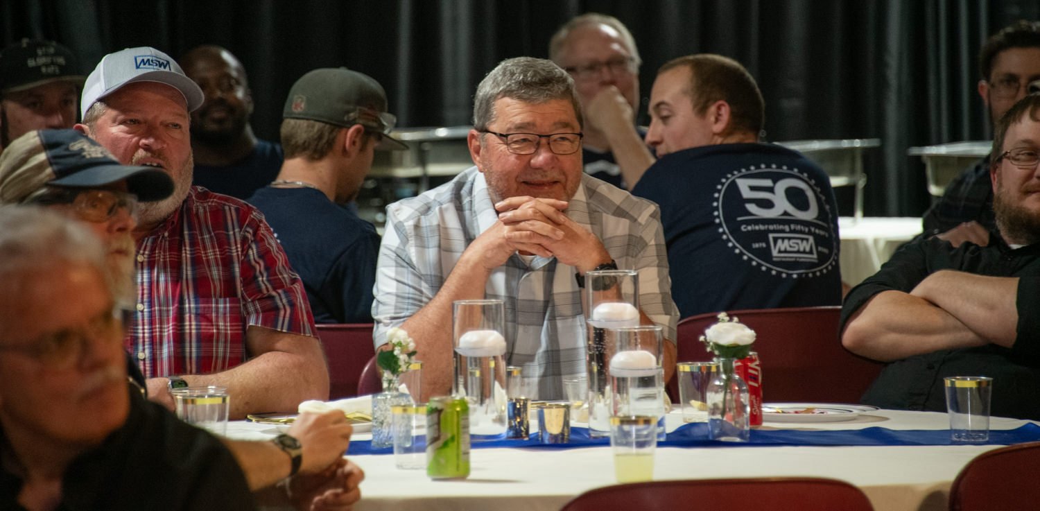 Group of men seated around a banquet table, drinks and flower centerpieces visible, man in a light plaid shirt rests his hands under his chin as others chat nearby.