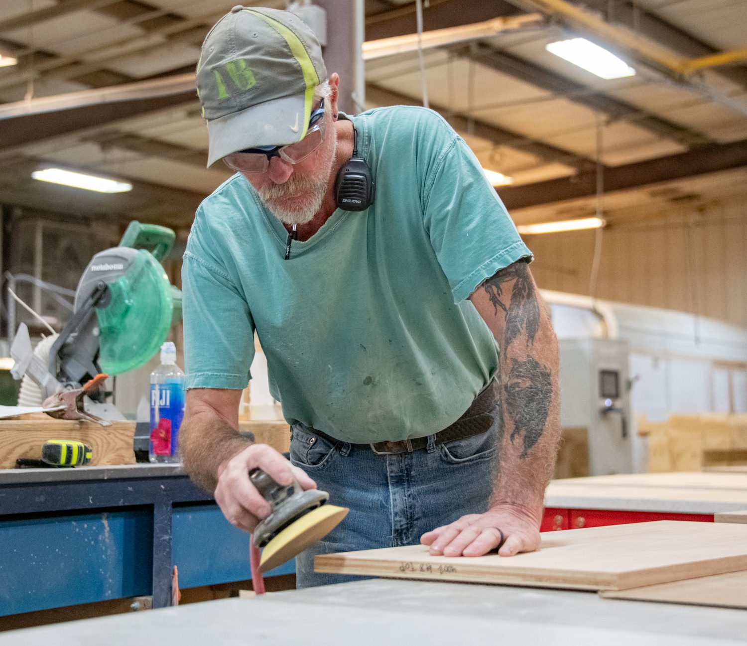 Woodworker wearing safety glasses and ear protection sands a wooden board with a handheld sander in a busy workshop.