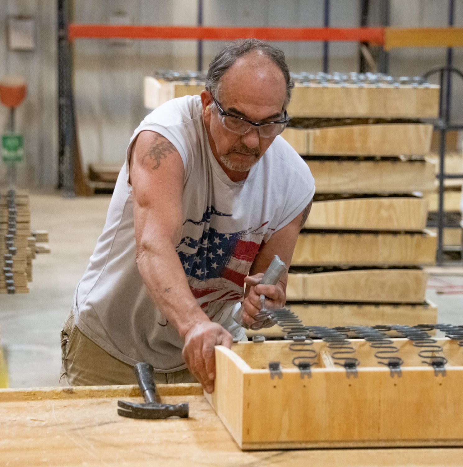 Man wearing safety glasses and an American flag t-shirt clamps wood pieces in a lumber workshop.