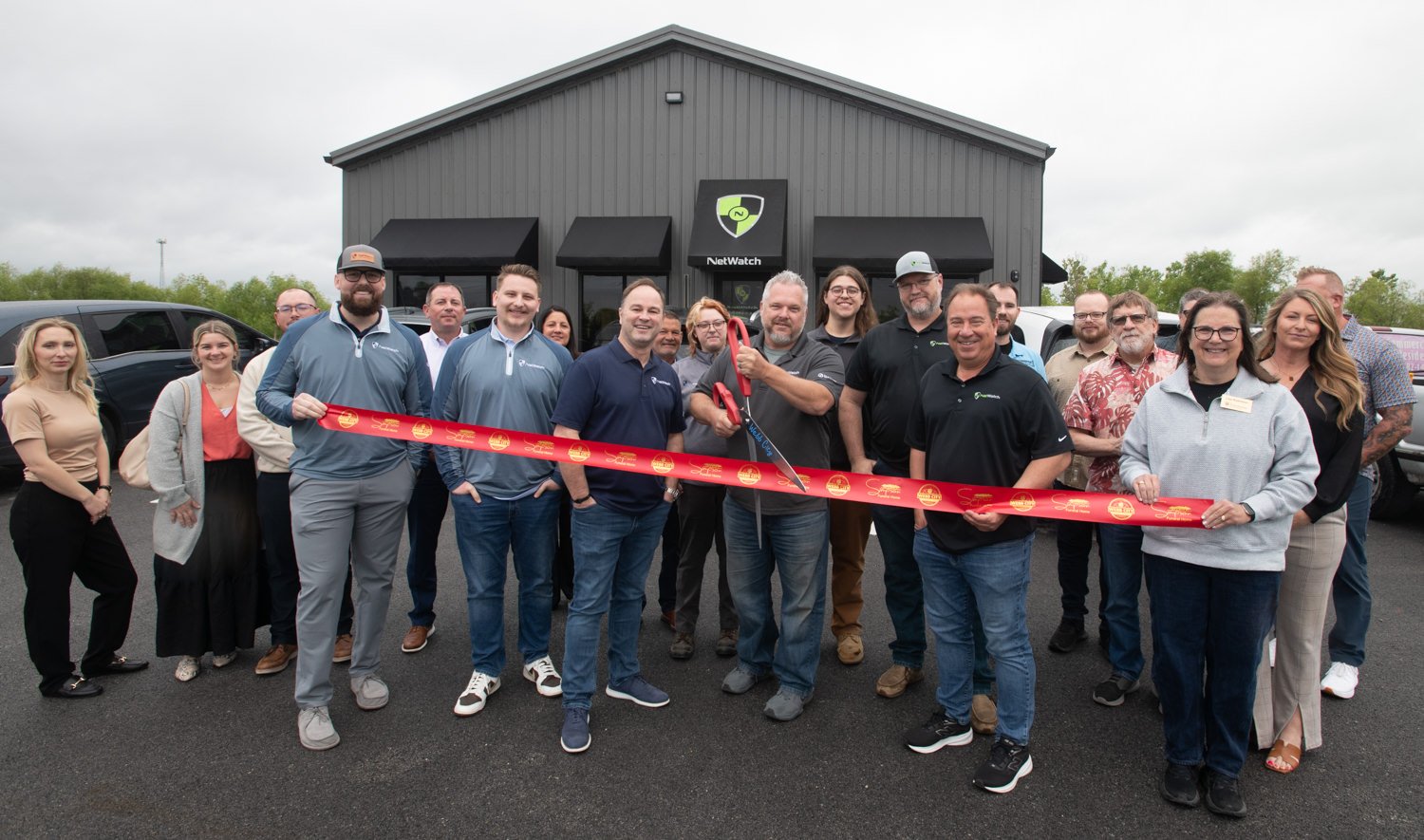 Group of people at a ribbon-cutting ceremony outside a gray industrial building with a NetWatch sign