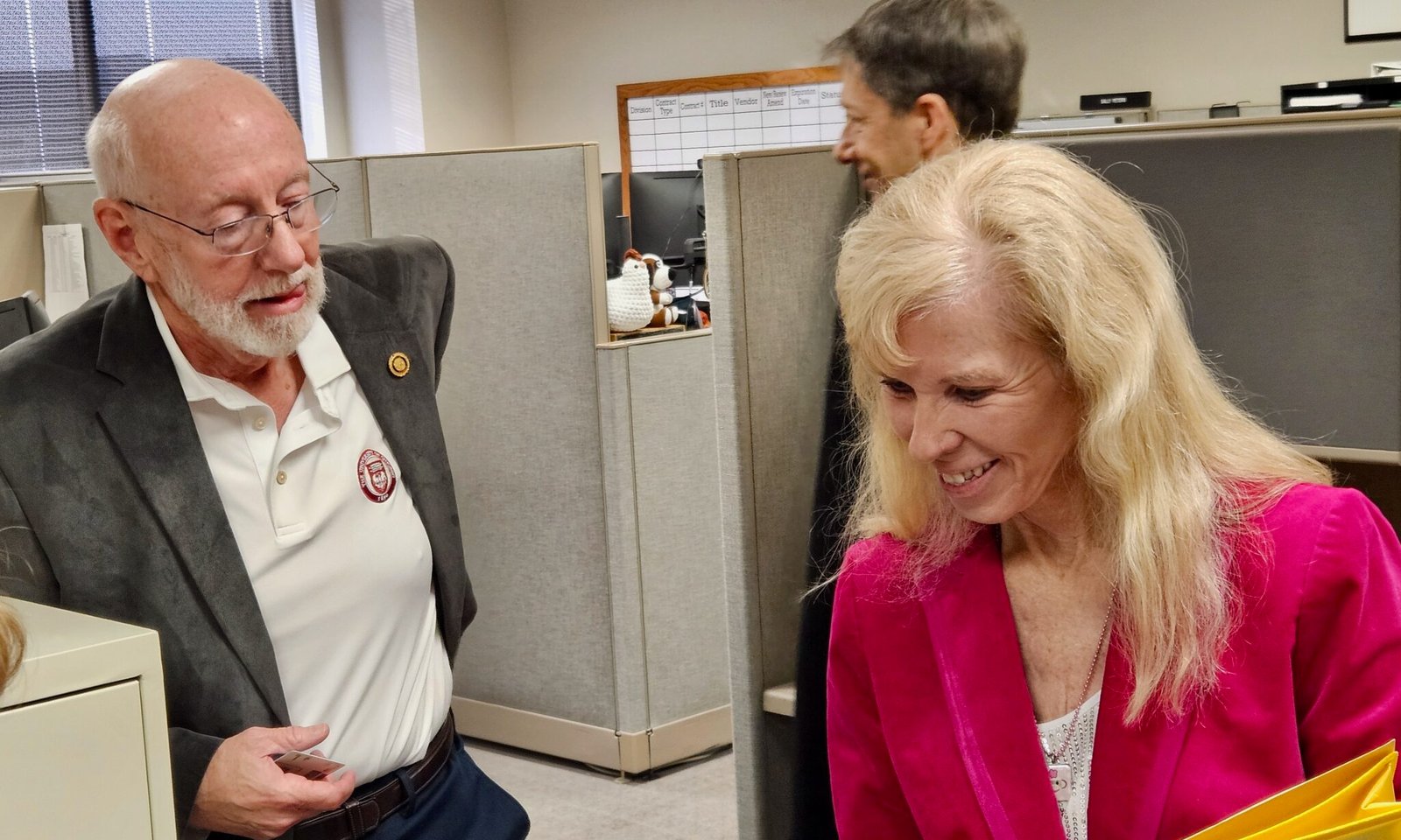Two coworkers in an office cubicle share a moment: man in a blazer and glasses handing a card to a smiling blonde woman in a pink jacket.