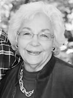 Smiling elderly woman with short white hair and glasses, wearing a dark jacket and a chain necklace, outdoors.