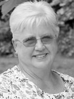 Portrait of an older woman with short white hair, glasses, and a floral blouse, smiling.