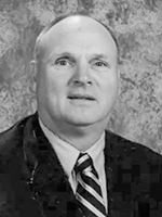 Middle-aged man in a dark suit and striped tie posing for a formal portrait against a textured gray background.