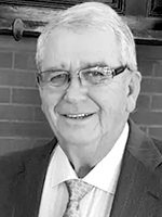 Smiling older man with glasses in a suit and tie, standing in front of a brick wall.