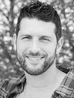 Smiling man with short dark hair and beard, wearing a plaid shirt, outdoors with blurred background.