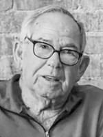 Portrait of an older man with gray hair and glasses, speaking in front of a brick wall.