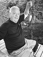 Older man sitting on a swing outdoors, smiling at the camera and raising one arm.