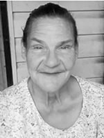 Smiling older woman with hair tied back, wearing a light patterned blouse, posed in front of a wooden slat wall.