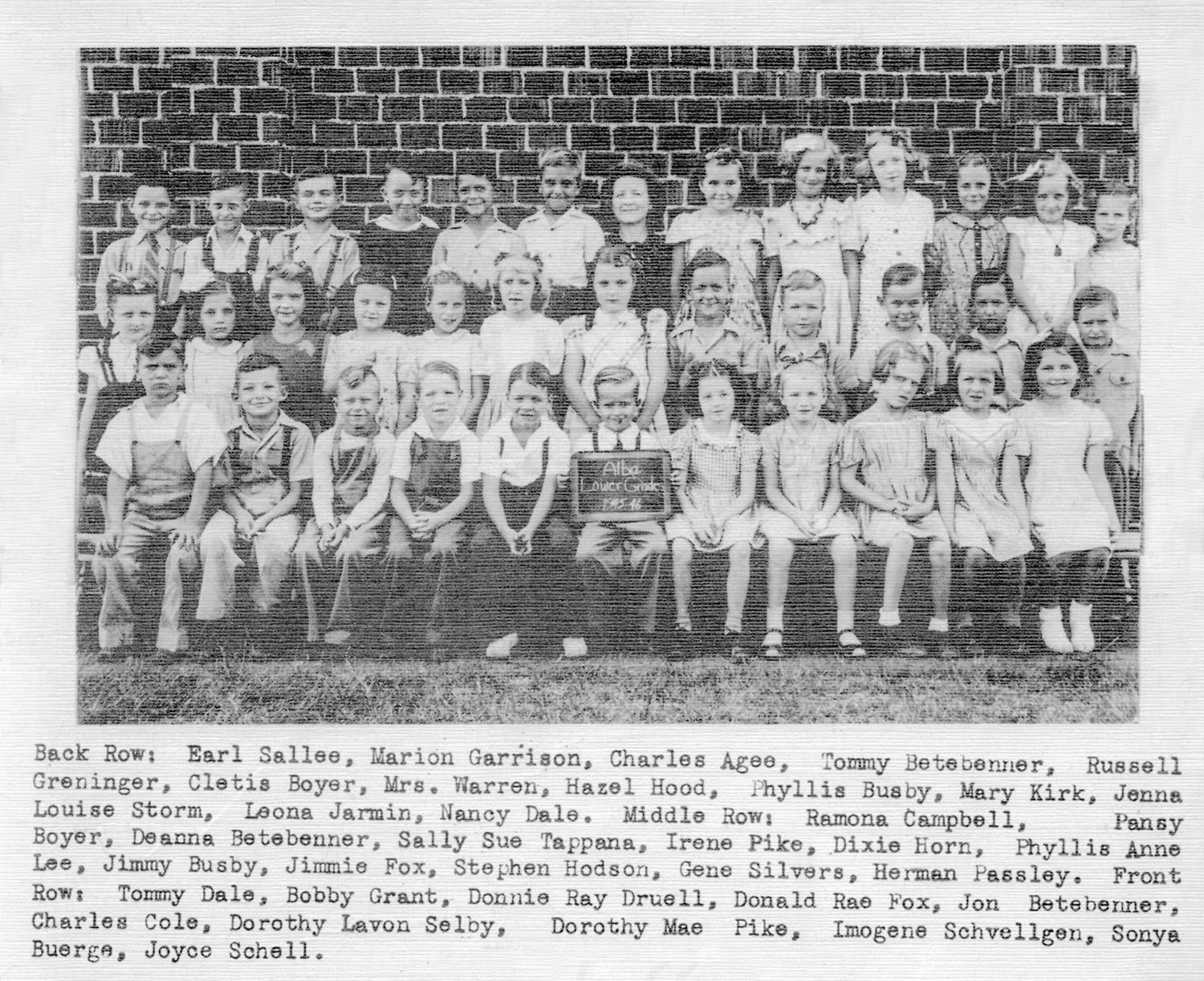 Group of many elementary school children posed for a class photo in front of a brick wall, arranged in four rows with a small plaque visible in the center front row.