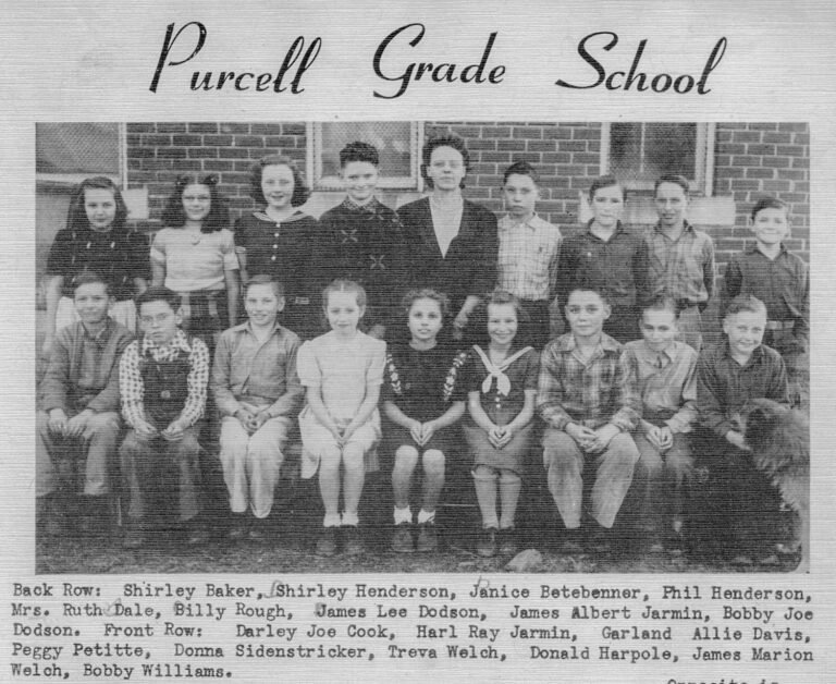Group of grade school students and teachers posing for a class photo in front of a brick building labeled Purcell Grade School.