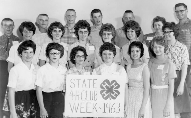 Group of teens and a few adults posing for a 4-H Club Week 1963 photo, holding a sign with the 4-H clover emblem.