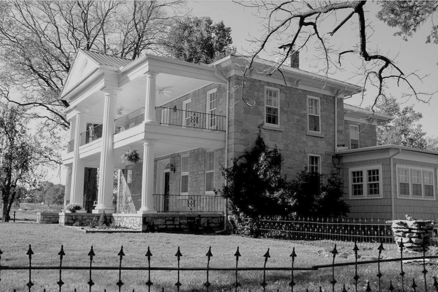 Large two-story house with prominent white columns on a wraparound porch, stone exterior, and a wrought-iron fence in the front yard.