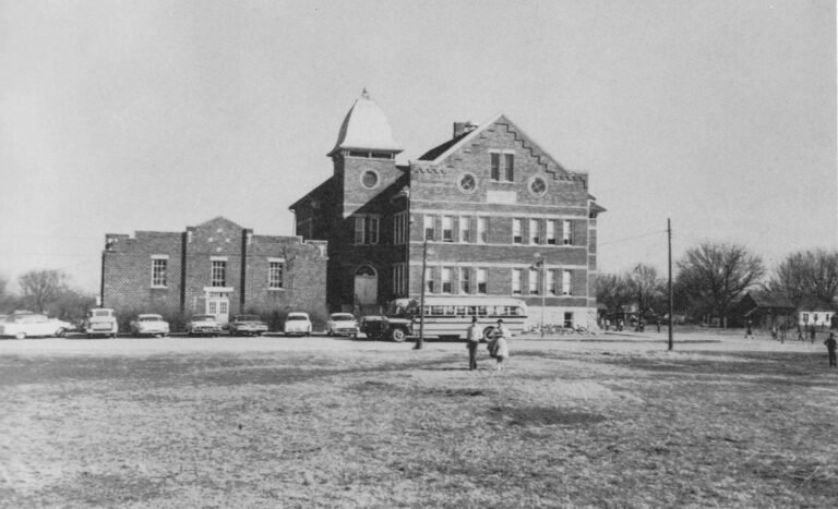 Historic brick school building with a tower and decorative gables, vintage cars parked along the curb, and a school bus in front; two children walk on the field in the foreground.