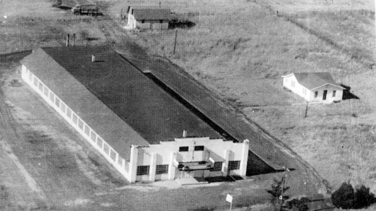 Aerial view of a long rectangular building with a sloped roof and a row of windows; a car is parked at the front entrance.