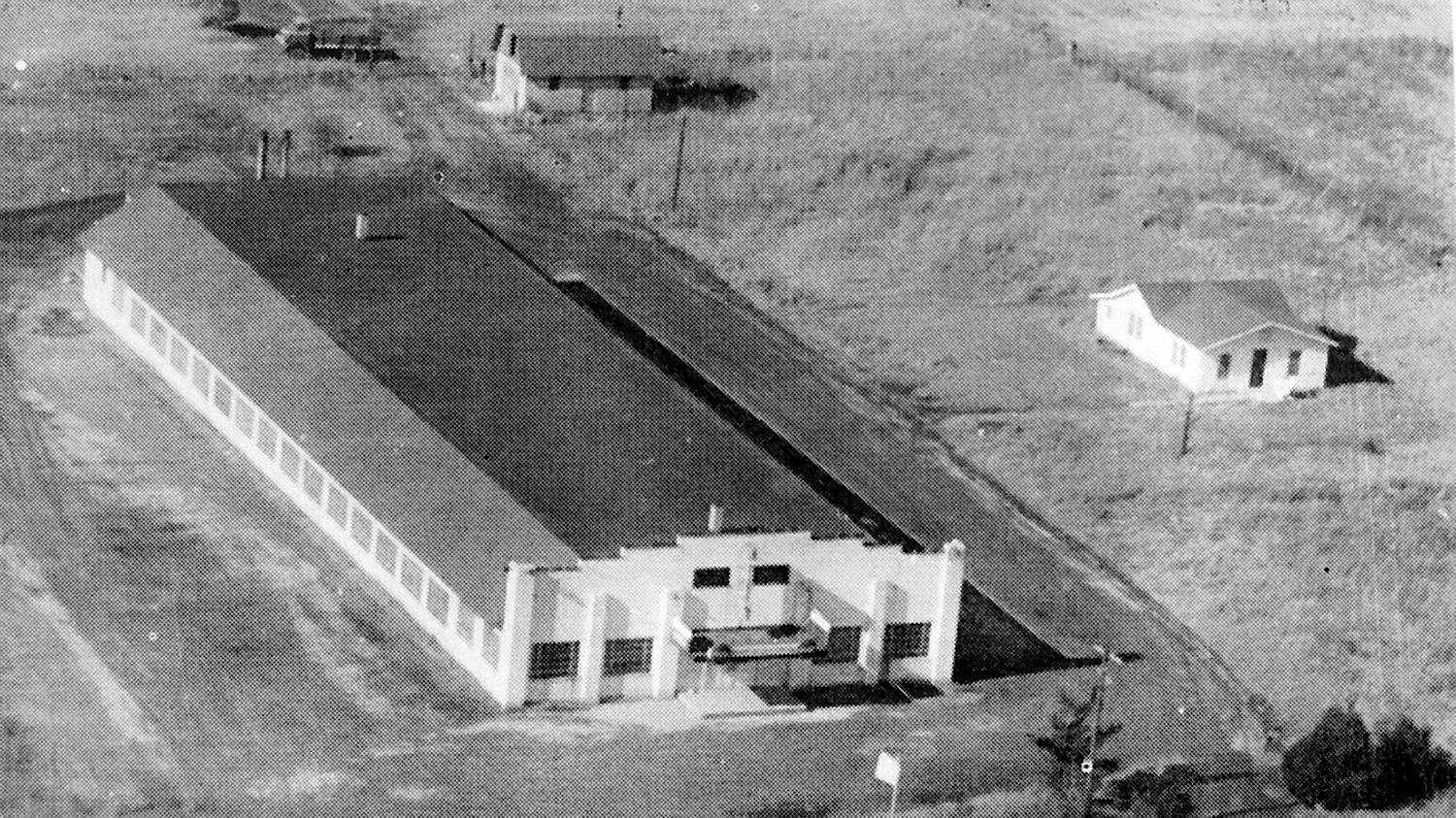 Aerial view of a long rectangular building with a sloped roof and a row of windows; a car is parked at the front entrance.