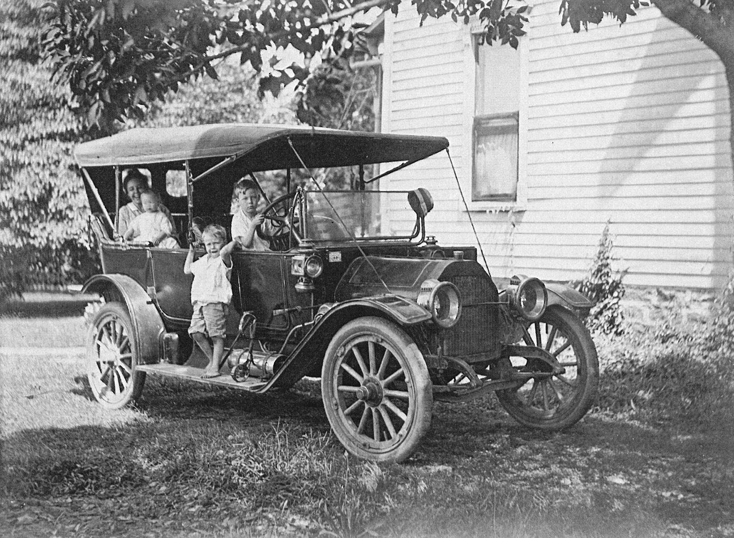 Vintage early 20th-century open-top automobile with several children posing beside it in a yard near a house.