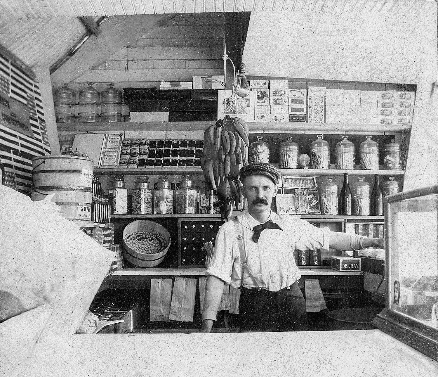Black-and-white image of a shopkeeper behind a small grocery counter, with bananas hanging overhead and jars on shelves behind him.