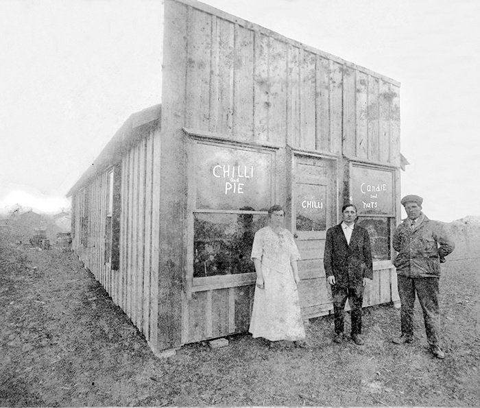 Three people stand outside a weathered wooden snack shop with chalked signs reading 'Chilli Pie' and 'Chilli' in a historical black-and-white photo.