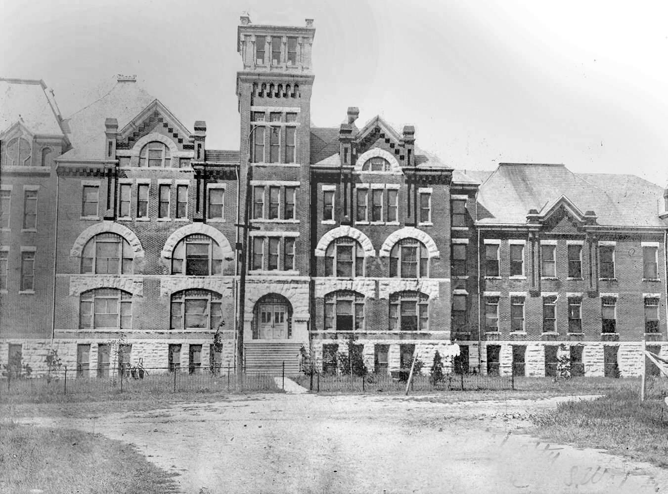 Historic brick building with arched first-floor windows, decorative stone trim, and a tall central tower, set behind a fence and dirt road.