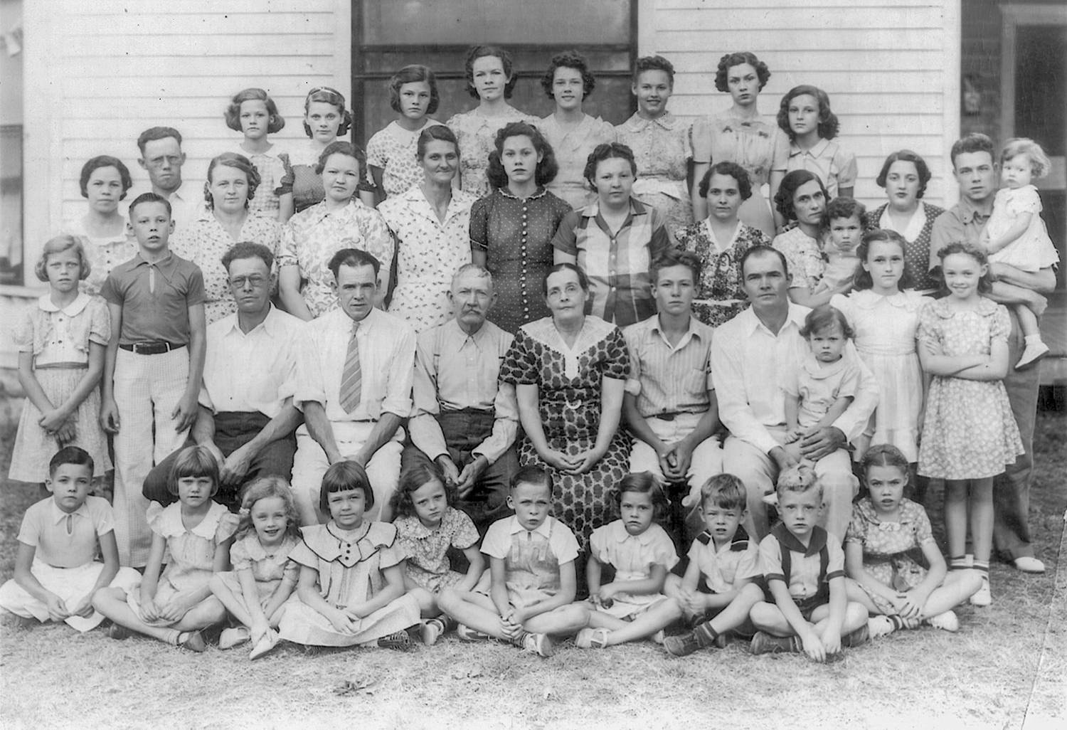 Large black-and-white group portrait of many adults and children in mid-20th-century clothing, posed outside a building.