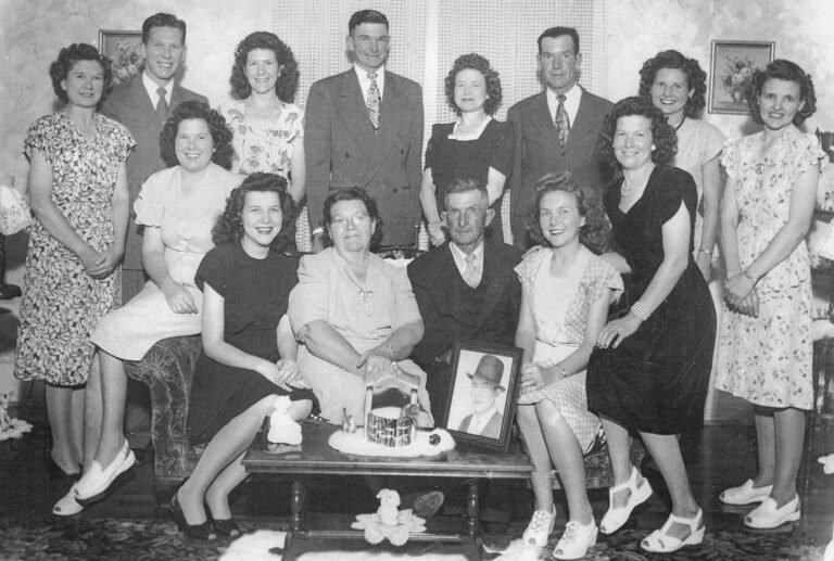 Group of adults and teens posing for a vintage family portrait in a living room, with a framed photo on the coffee table and a cake centerpiece in front of them.